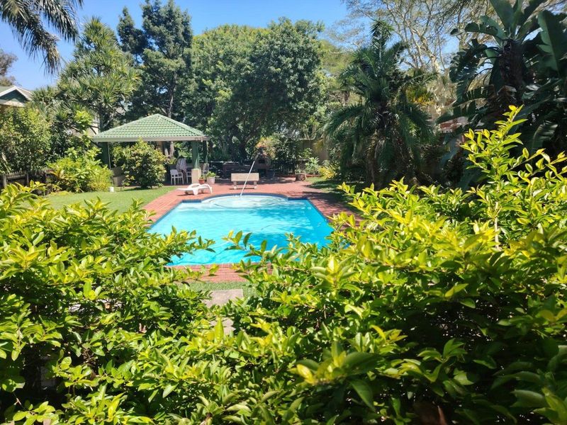 A well-maintained outdoor pool area featuring a swimming pool, brick paved surround, and a shaded gazebo set within a lush, landscaped garden.