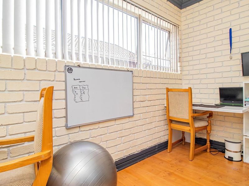 A functional home office space featuring painted brick walls, laminate flooring, and large windows with vertical blinds, providing ample natural light.