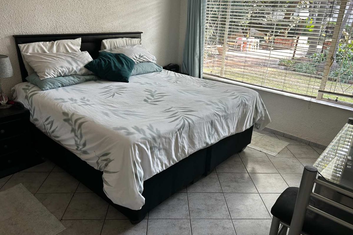 A bedroom featuring tiled flooring and large windows with blinds, offering natural light and a view of the outdoor garden area.