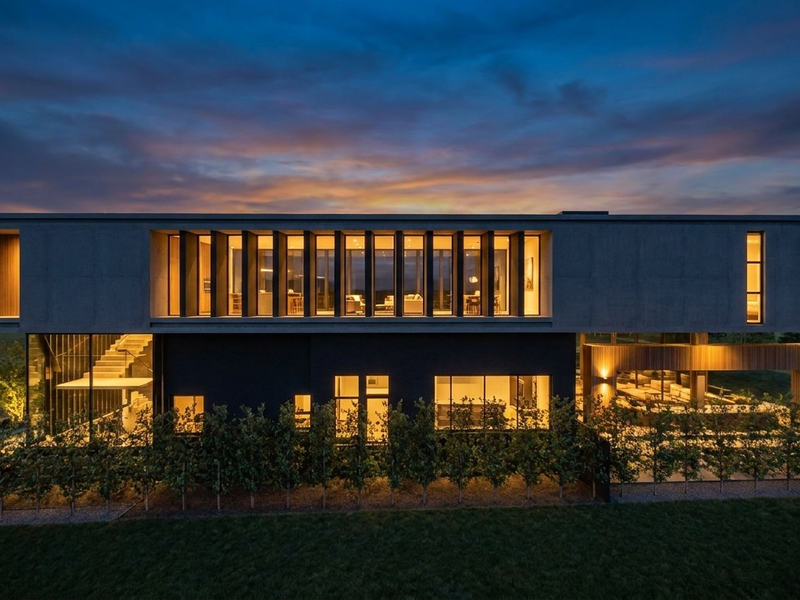 A striking modern architectural residence at dusk, featuring a concrete upper level, expansive glazing, integrated lighting, and a landscaped rear garden area.