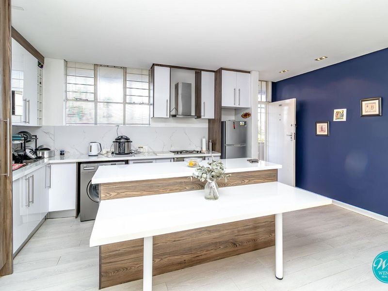 Modern kitchen featuring sleek white and wood-grain cabinetry, stone countertops, integrated appliances, and a functional breakfast island, complemented by a striking navy accent wall.