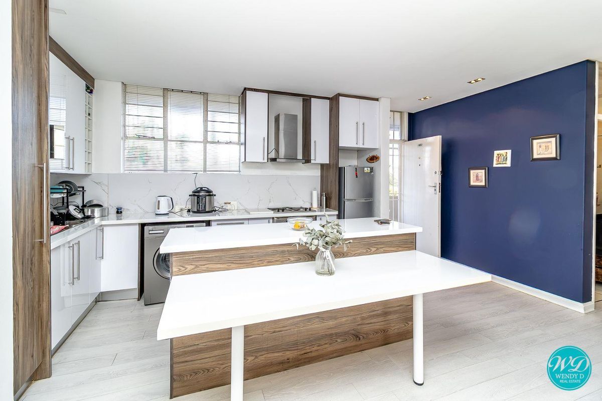 Modern kitchen featuring sleek white and wood-grain cabinetry, stone countertops, integrated appliances, and a functional breakfast island, complemented by a striking navy accent wall.