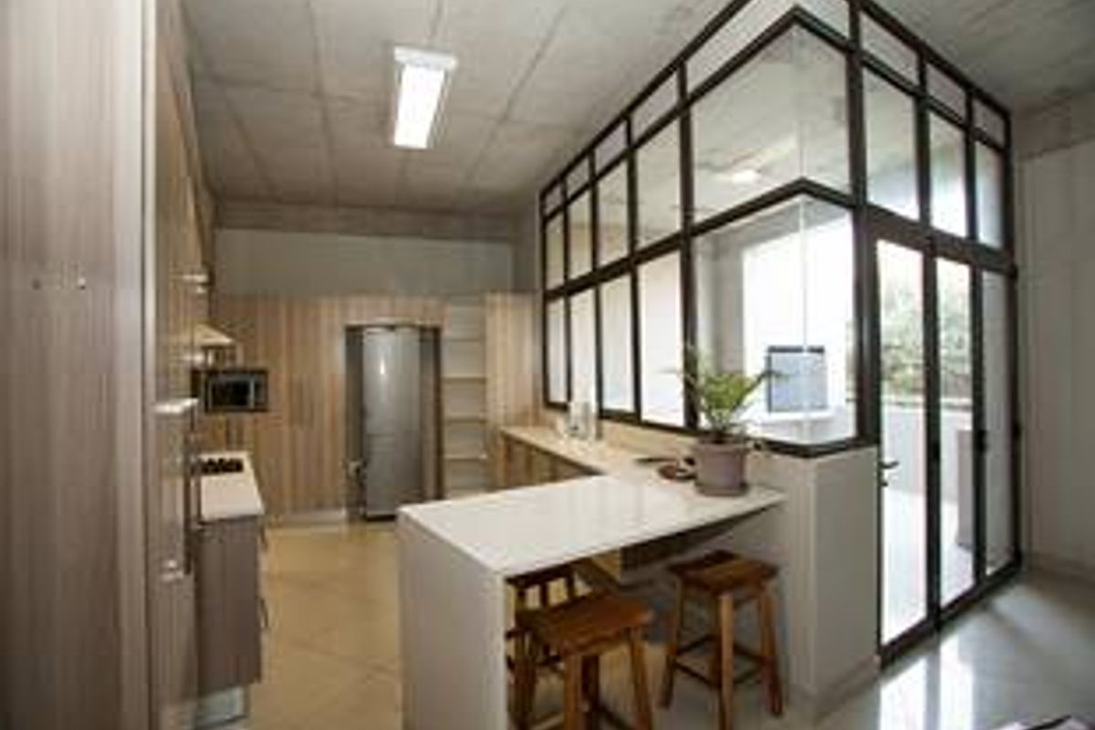 Modern kitchen featuring light wood cabinetry, stone countertops, a breakfast bar, and a striking industrial-style glass partition wall with exposed concrete ceilings.