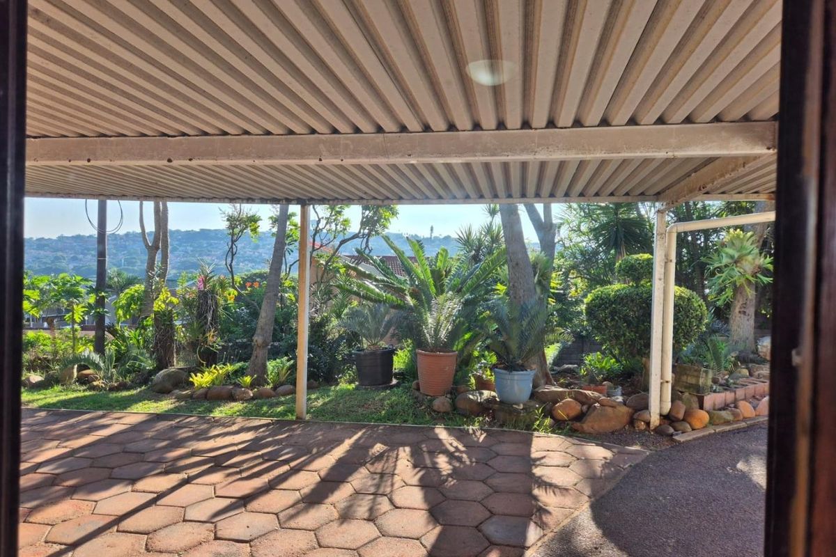 A covered outdoor patio area featuring paved flooring and a corrugated metal roof, overlooking a landscaped garden with mature plants.