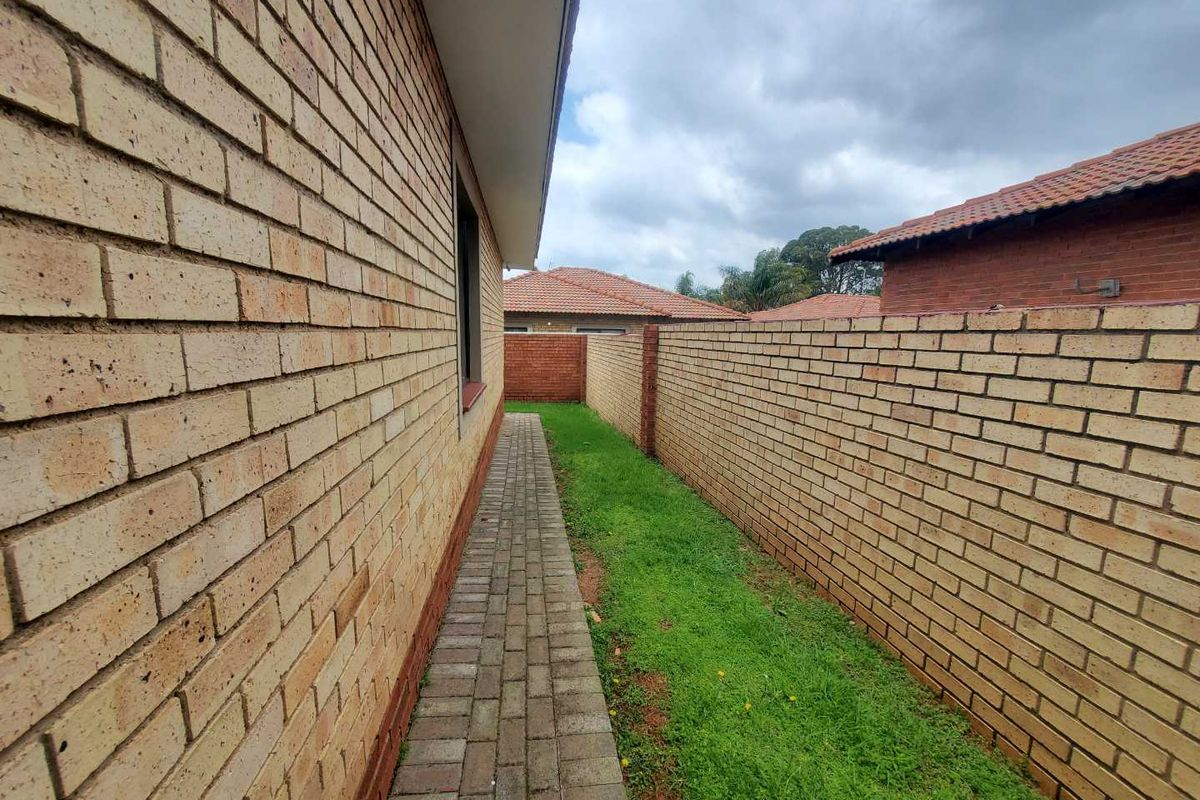 A side exterior walkway featuring paved pathing alongside the brick house wall and a perimeter boundary wall with a grass verge.