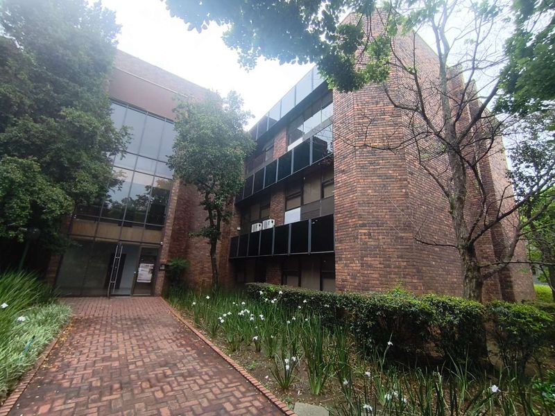 A multi-story office building featuring a classic brick facade, large glass entrance, and a paved walkway surrounded by established garden landscaping.