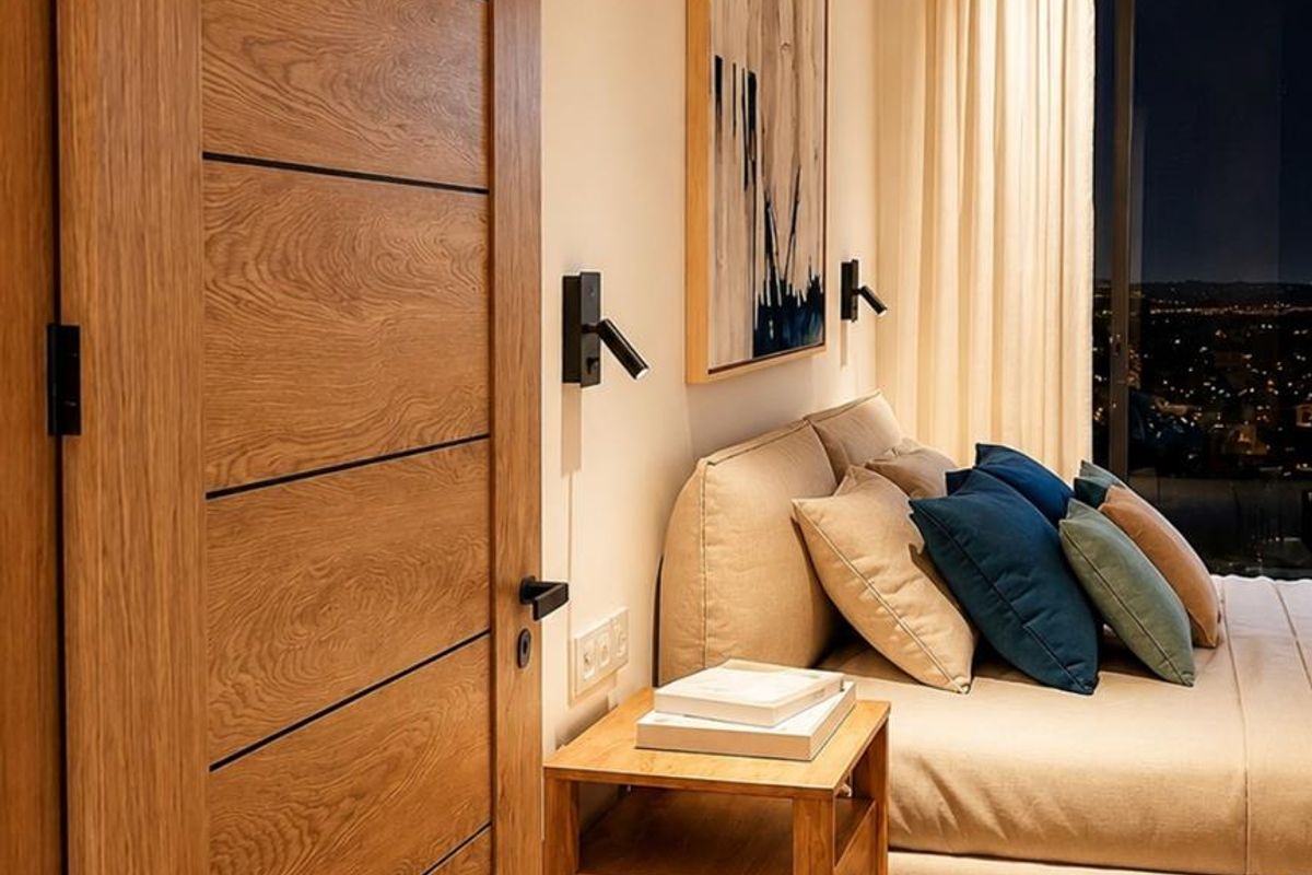 A modern bedroom featuring a wooden door, recessed lighting, and large windows offering a night view of the city.