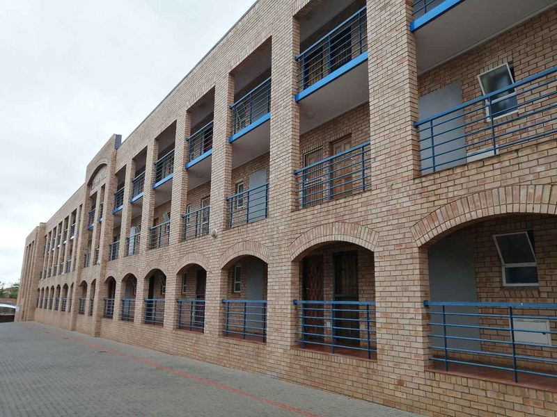 A multi-story apartment building facade featuring face brick construction, blue metal balustrades, and arched architectural details with a paved driveway.