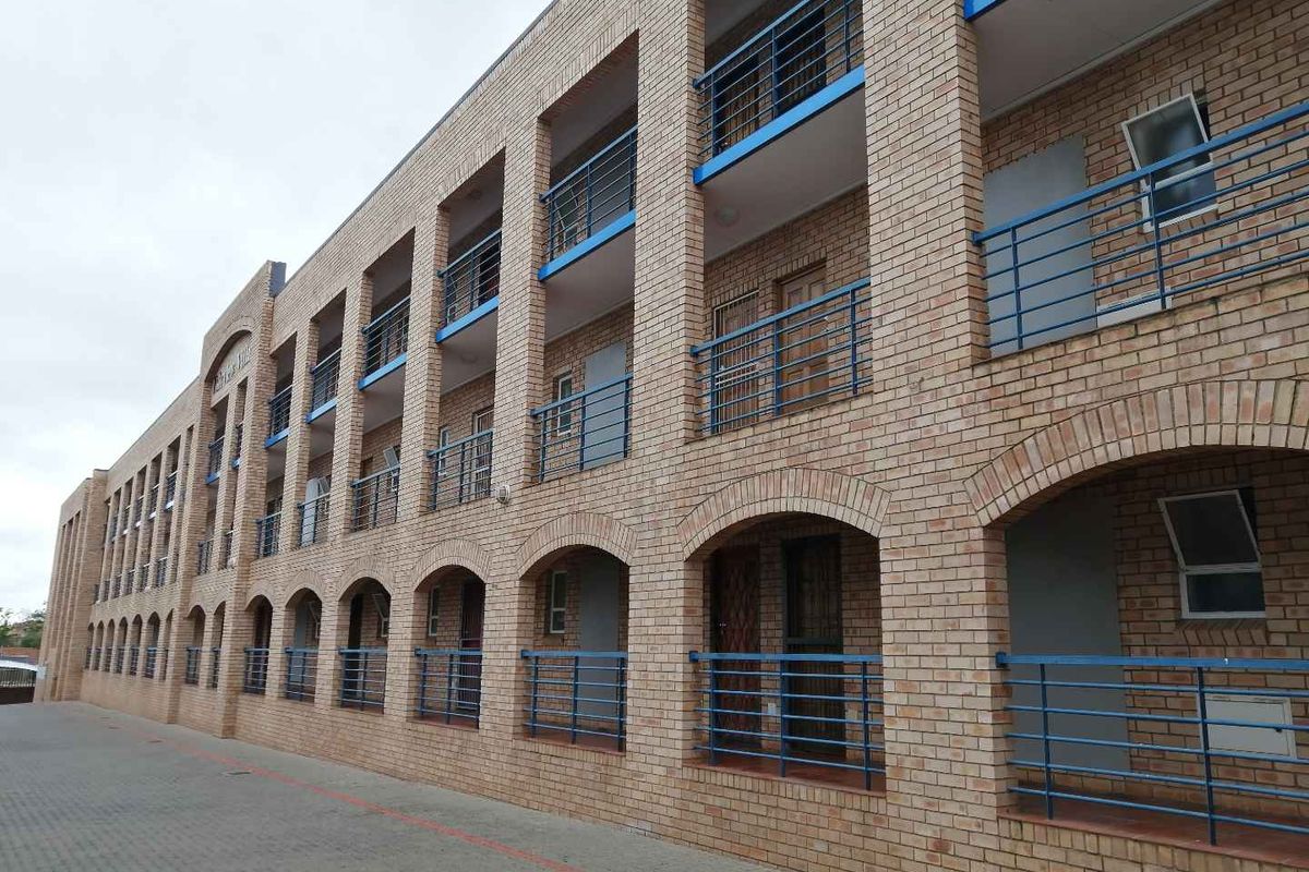 A multi-story apartment building facade featuring face brick construction, blue metal balustrades, and arched architectural details with a paved driveway.