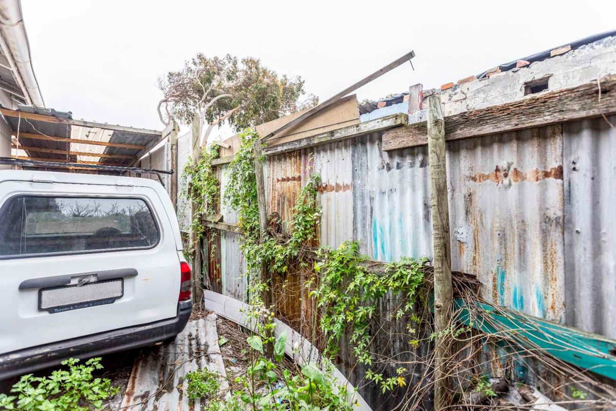 A side exterior area featuring a heavily weathered corrugated metal fence, overgrown vegetation, and signs of significant structural deterioration.