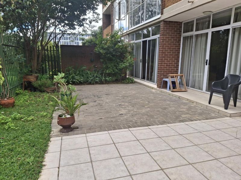 A private rear patio area featuring a combination of brick paving and tiled flooring, directly accessible from the apartment through sliding glass doors.