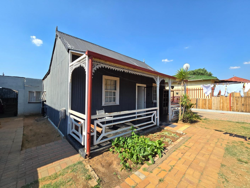 A charming cottage featuring corrugated iron cladding, a classic veranda with decorative fretwork, and a paved front garden area.