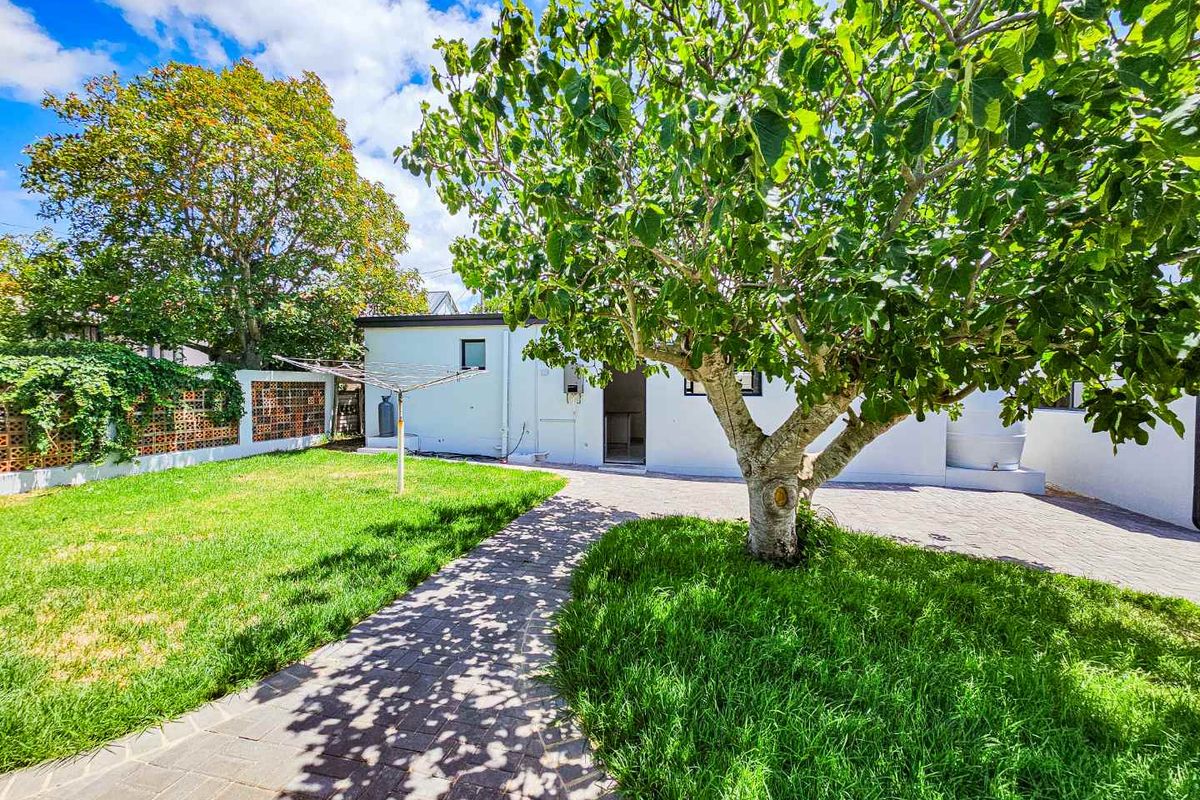 A private rear garden featuring a paved walkway, established tree, and a white exterior wall with access to the main dwelling.