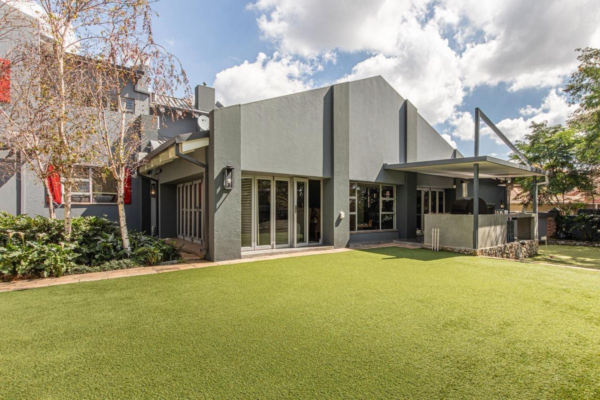 Rear view of the property featuring a modern grey facade, covered patio with built-in braai, and a well-maintained artificial lawn garden area.