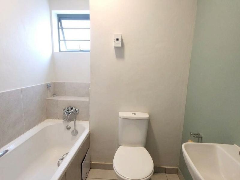 A functional bathroom featuring a bathtub, toilet, and wall-mounted washbasin with neutral tiling and a louver window for ventilation.