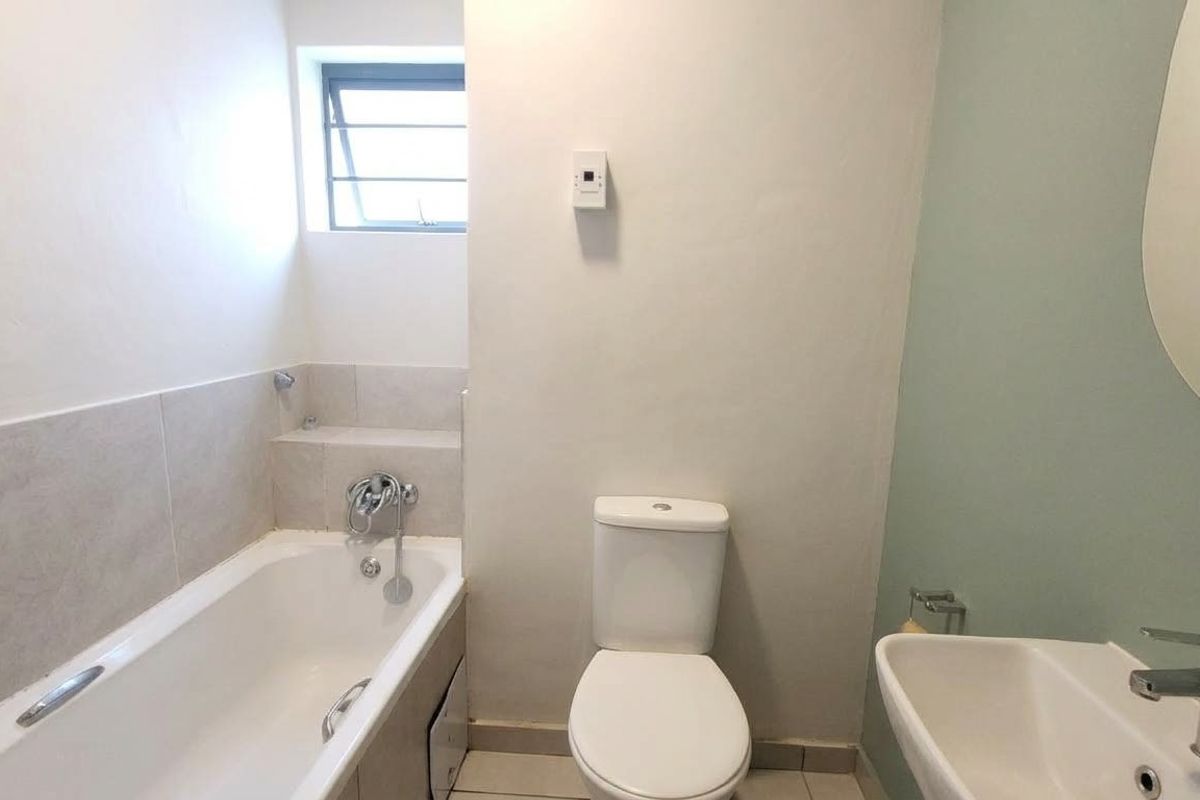 A functional bathroom featuring a bathtub, toilet, and wall-mounted washbasin with neutral tiling and a louver window for ventilation.
