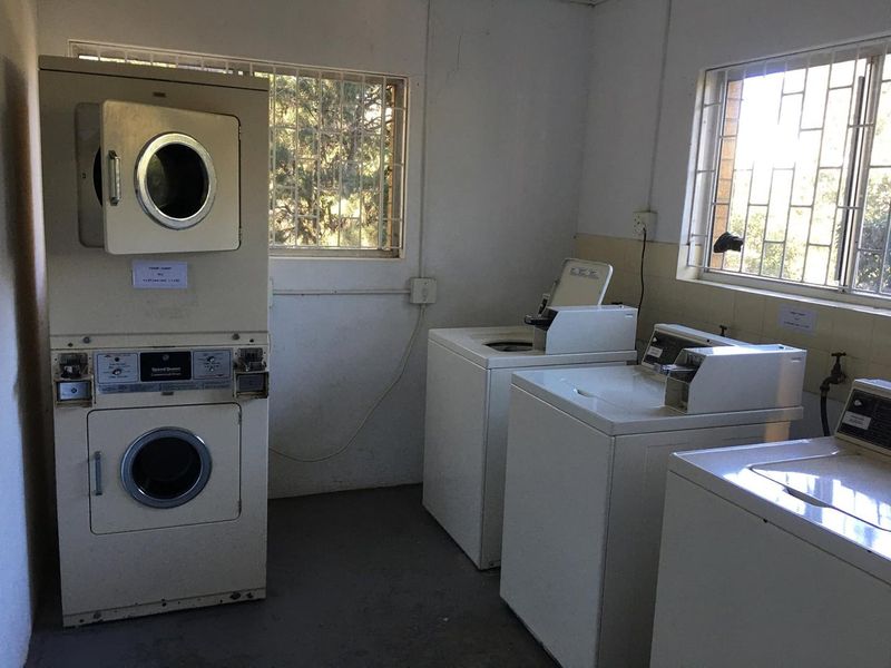 A functional communal laundry room featuring multiple washing machines and a stacked dryer unit, equipped with security bars on the windows.