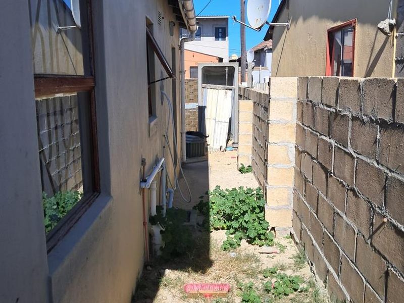 A narrow side or rear exterior passage featuring a concrete block boundary wall, external plumbing, and unpaved ground area.
