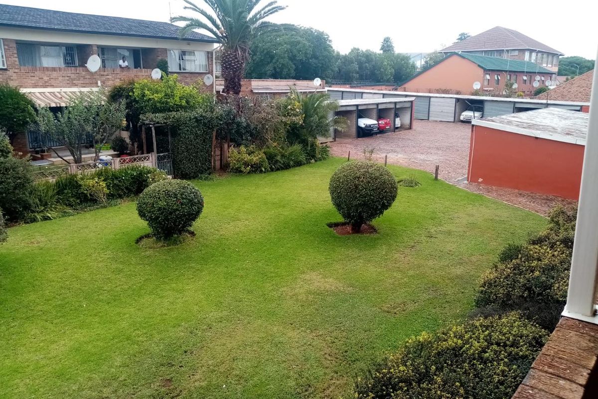 View of a well-maintained communal garden area with established landscaping, brick apartment building, and covered parking bays.