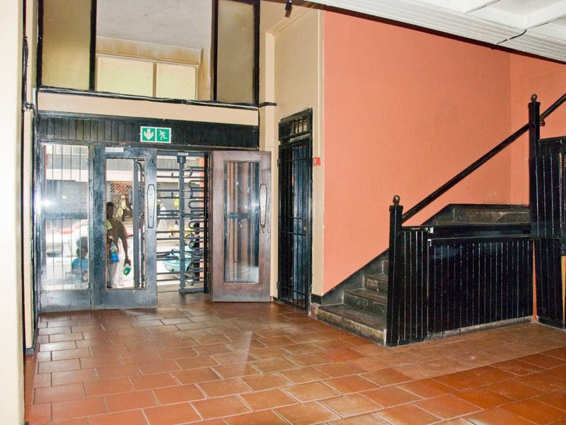 A spacious apartment building entrance lobby featuring terracotta tiled flooring, a security turnstile, and a staircase with a wooden balustrade.