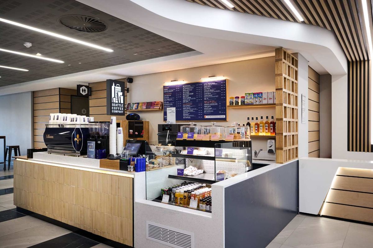 A modern, well-maintained coffee service counter featuring integrated cabinetry, professional display shelving, and contemporary recessed ceiling lighting within an office environment.