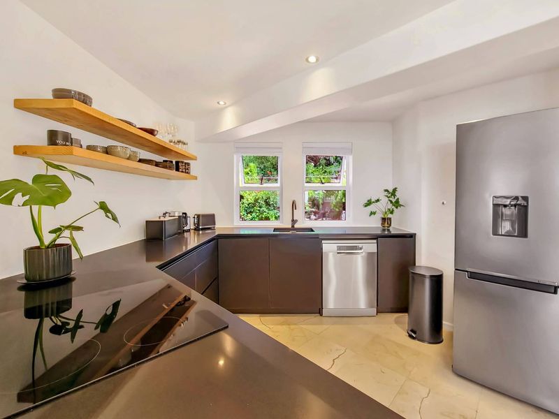 Modern kitchen featuring dark cabinetry, stone countertops, integrated hob, stainless steel dishwasher, and floating timber shelving with views to the garden.