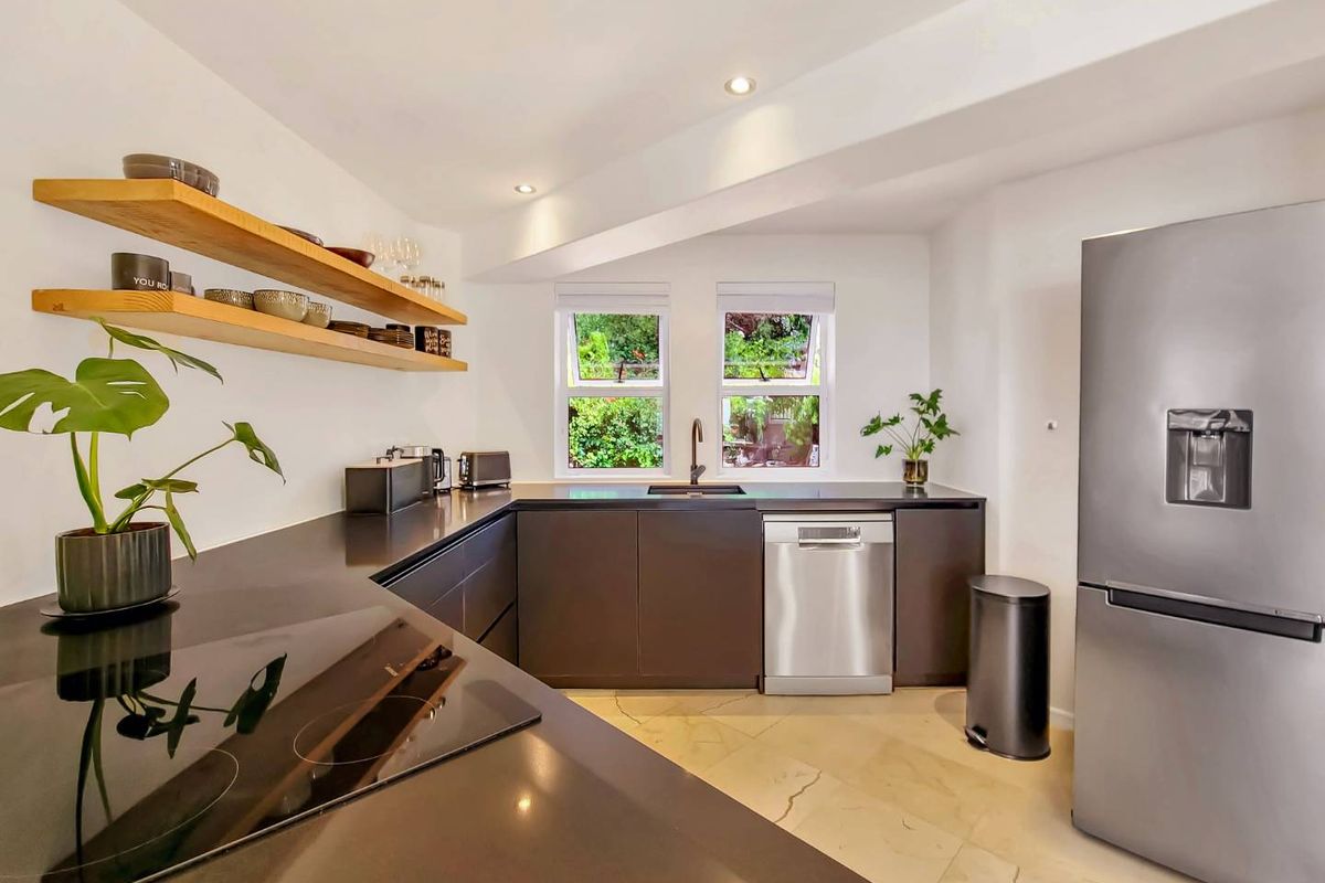Modern kitchen featuring dark cabinetry, stone countertops, integrated hob, stainless steel dishwasher, and floating timber shelving with views to the garden.