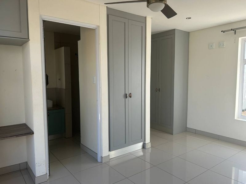 A bright, empty bedroom featuring neutral tiled flooring, built-in grey cabinetry, a ceiling fan, and an en-suite bathroom entrance.