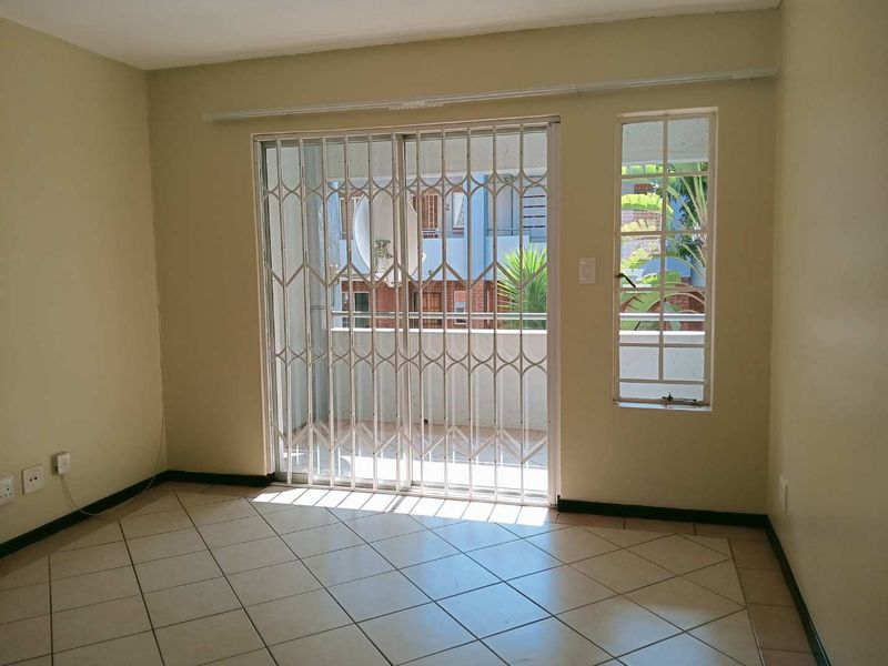An empty living room featuring tiled flooring, a sliding security gate leading to a balcony, and a fixed window providing natural light.