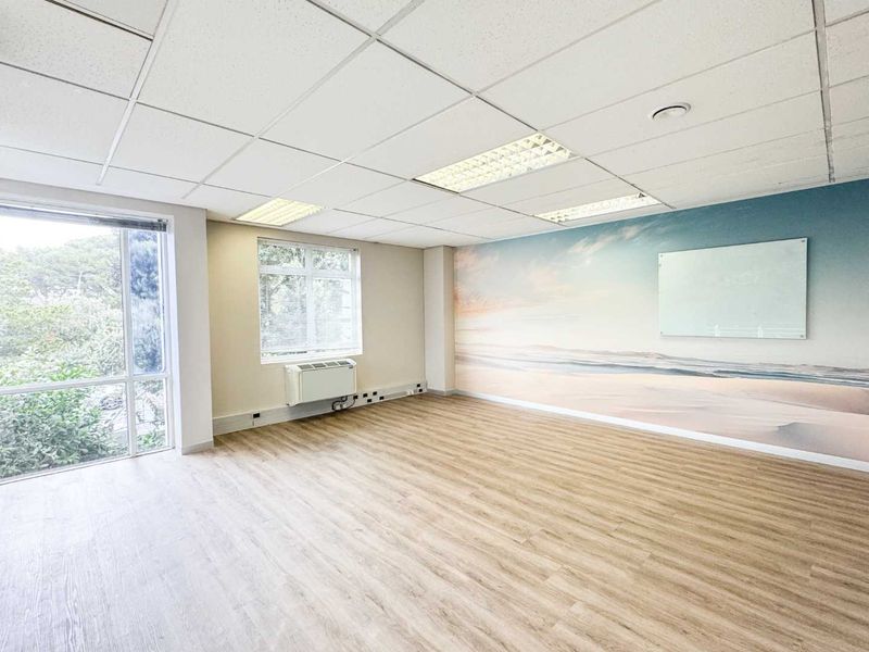 A bright, vacant office space featuring wood-look vinyl flooring, a suspended ceiling with recessed lighting, a wall-mounted HVAC unit, and a large window.