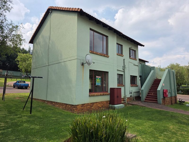 Rear view of a two-story townhouse featuring a tiled roof, external staircase, and brick base, set within a communal garden area.
