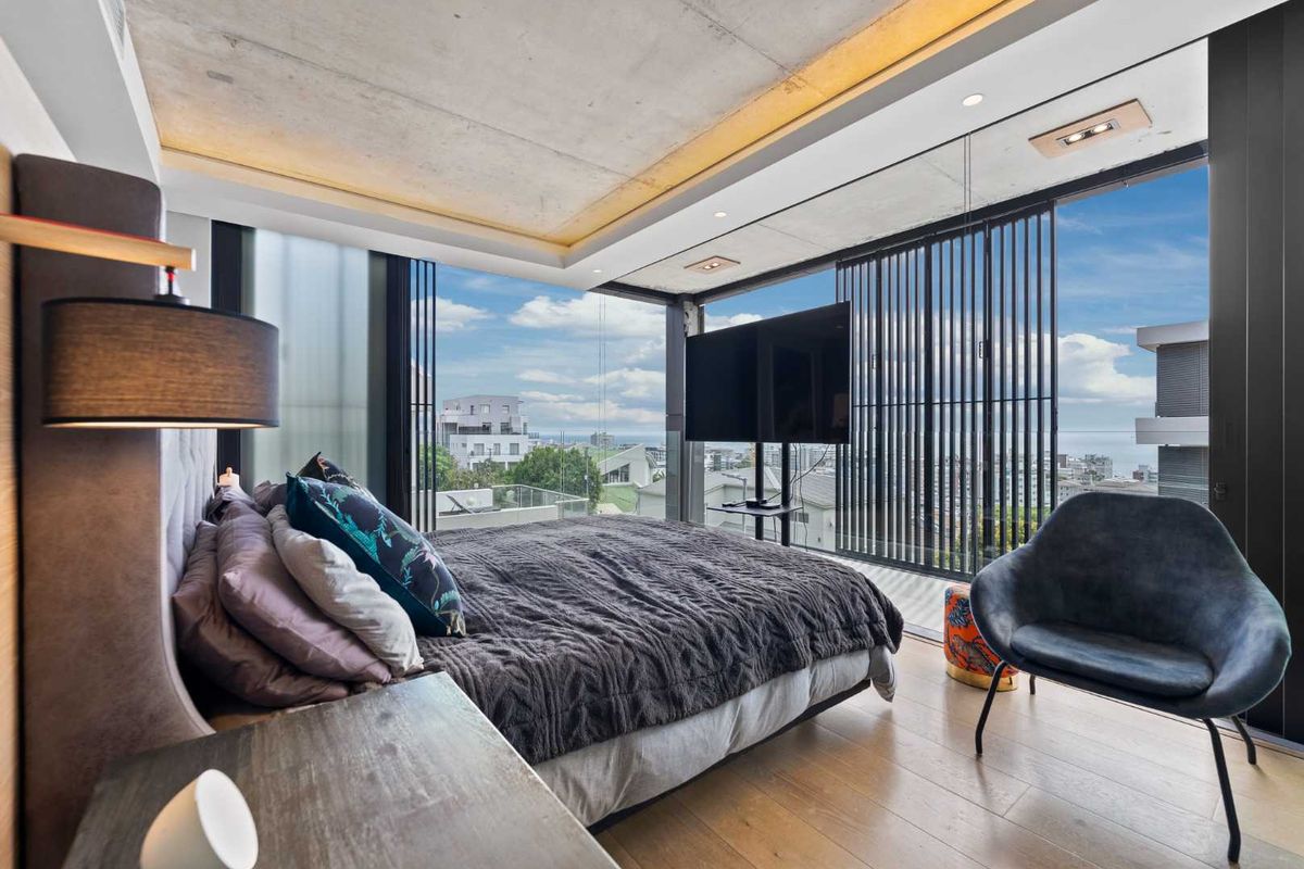 Modern bedroom featuring an exposed concrete ceiling, floor-to-ceiling windows with integrated metal shutters, and high-quality timber flooring with expansive urban and coastal views.