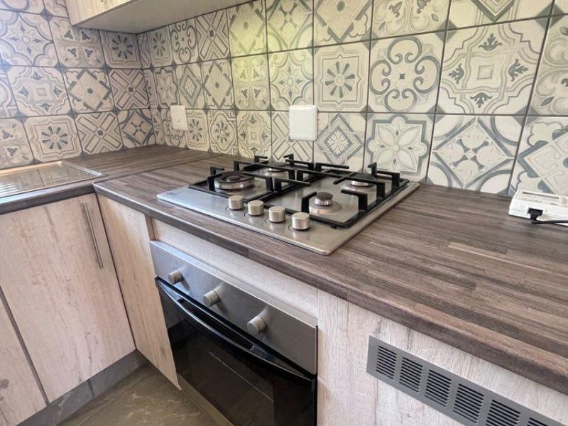 A compact, modern kitchen featuring light wood-grain cabinetry, dark laminate countertops, a gas hob, built-in oven, and decorative patterned tile backsplash.