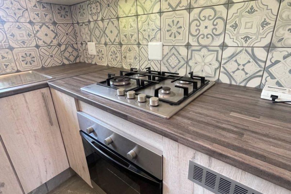 A compact, modern kitchen featuring light wood-grain cabinetry, dark laminate countertops, a gas hob, built-in oven, and decorative patterned tile backsplash.