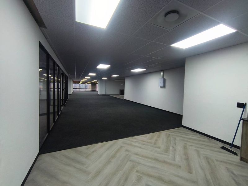 Spacious open-plan office area featuring a mix of carpet and herringbone vinyl flooring, recessed ceiling lights, and modern glass partitioning.