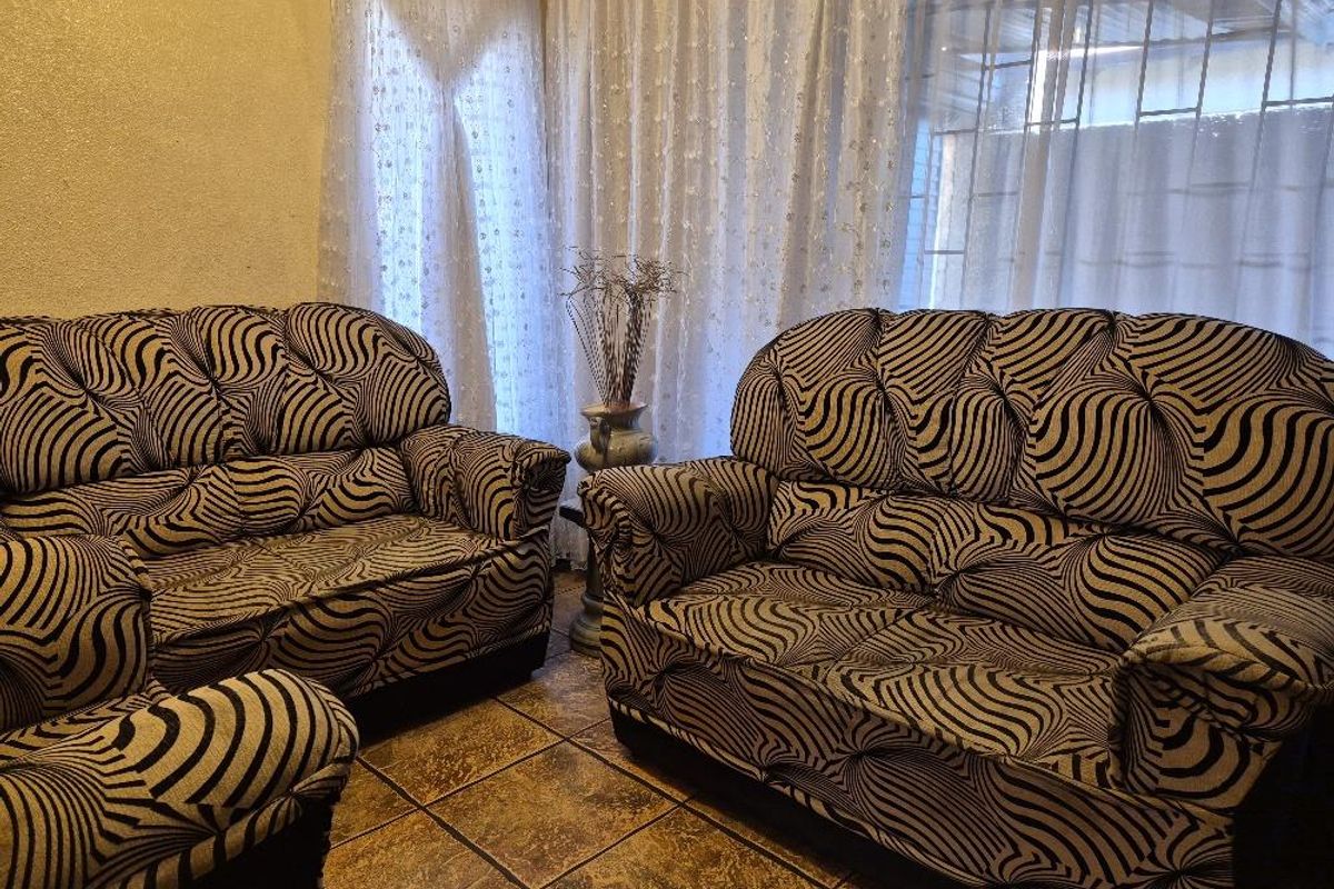 A living room area featuring tiled flooring and large windows covered by sheer curtains, providing natural light to the space.