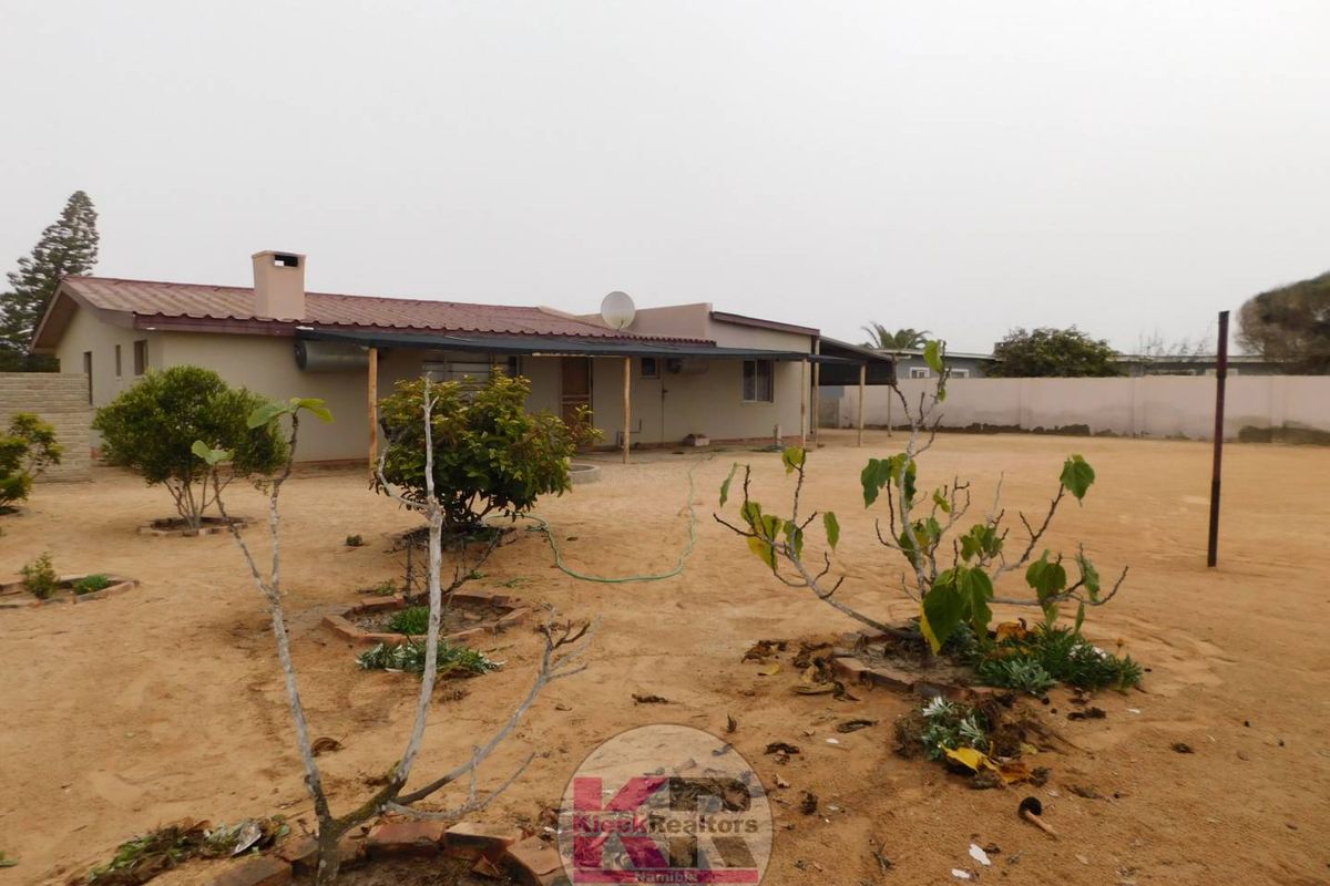 Rear view of the property featuring a covered patio, metal roof, and a large, sandy backyard enclosed by a boundary wall.