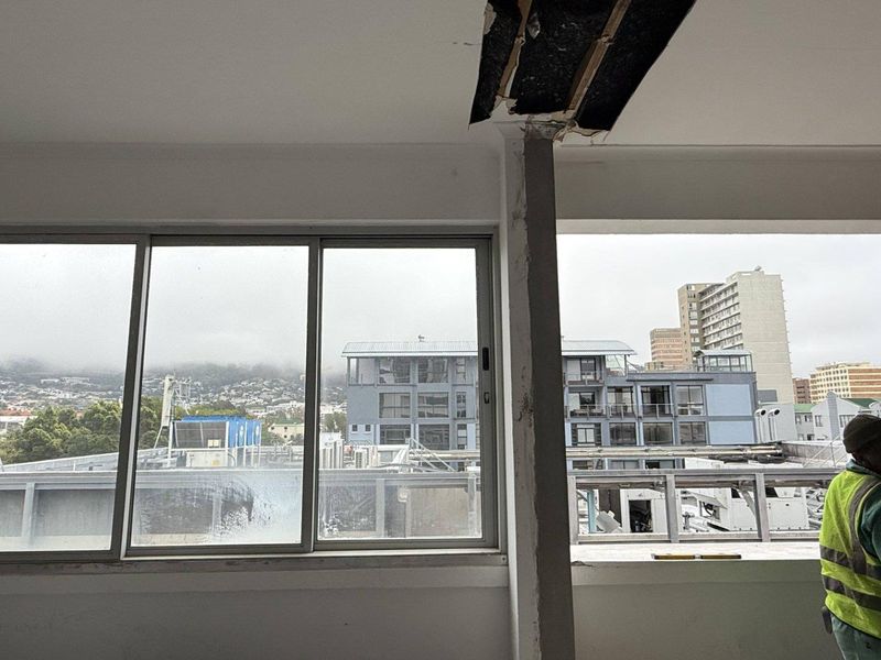 An office space currently undergoing renovation, featuring large sliding windows with urban views and exposed ceiling and wall sections.