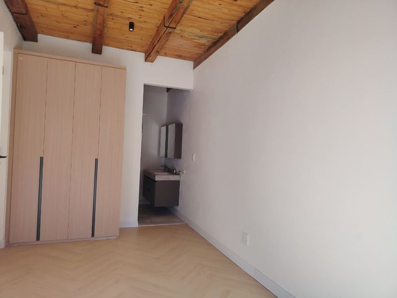 A bright bedroom featuring exposed timber ceiling beams, modern herringbone flooring, and a built-in wardrobe with direct access to an en-suite bathroom.