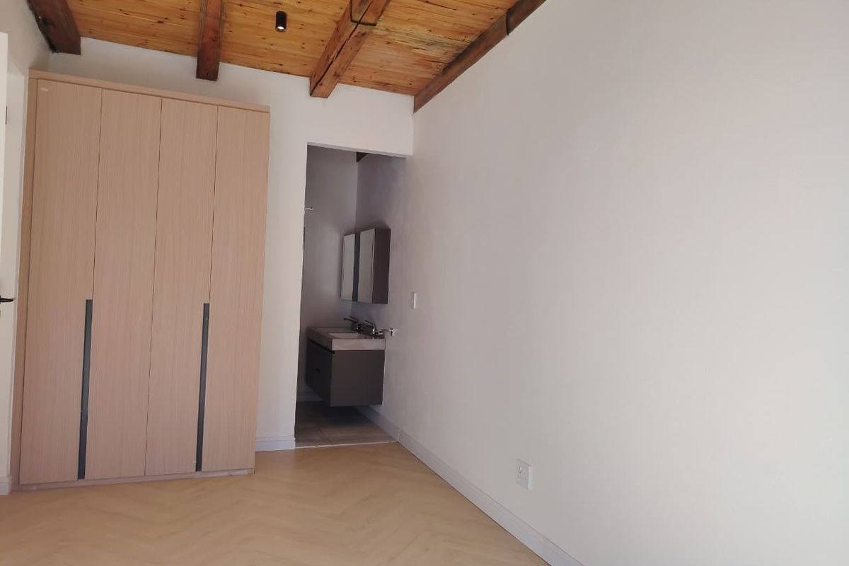 A bright bedroom featuring exposed timber ceiling beams, modern herringbone flooring, and a built-in wardrobe with direct access to an en-suite bathroom.