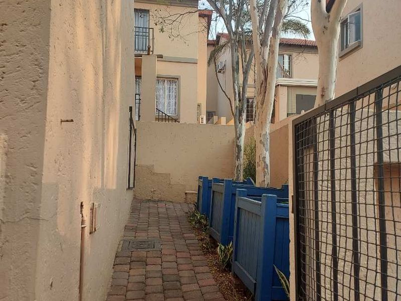 A narrow paved side walkway leading to the rear of the apartment complex, featuring a metal security gate and established trees.
