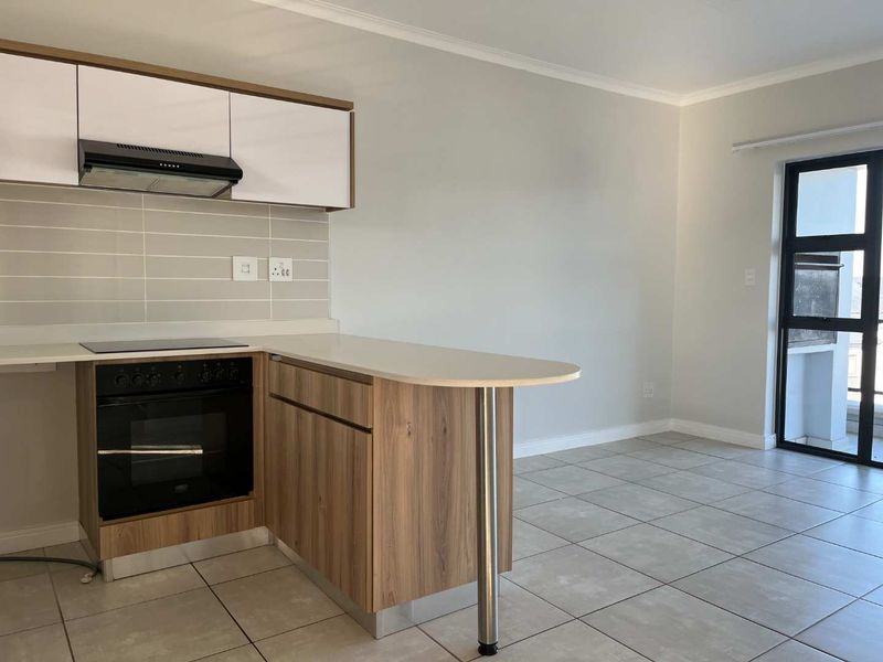 Modern open-plan kitchen featuring wood-finish cabinetry, integrated appliances, and a breakfast bar, flowing into a bright living area with tiled floors.