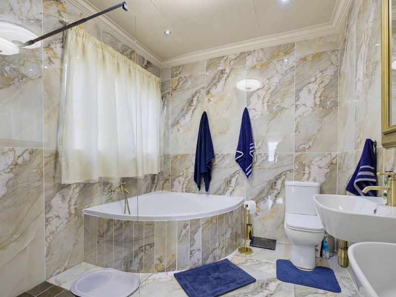 A well-maintained bathroom featuring marble-effect wall tiling, a corner bathtub, a pedestal sink, and modern brass fixtures.
