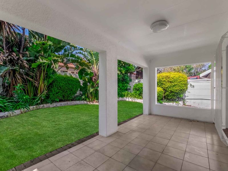 A clean, covered patio area featuring tiled flooring and concrete pillars, overlooking a well-maintained private garden with lush greenery.