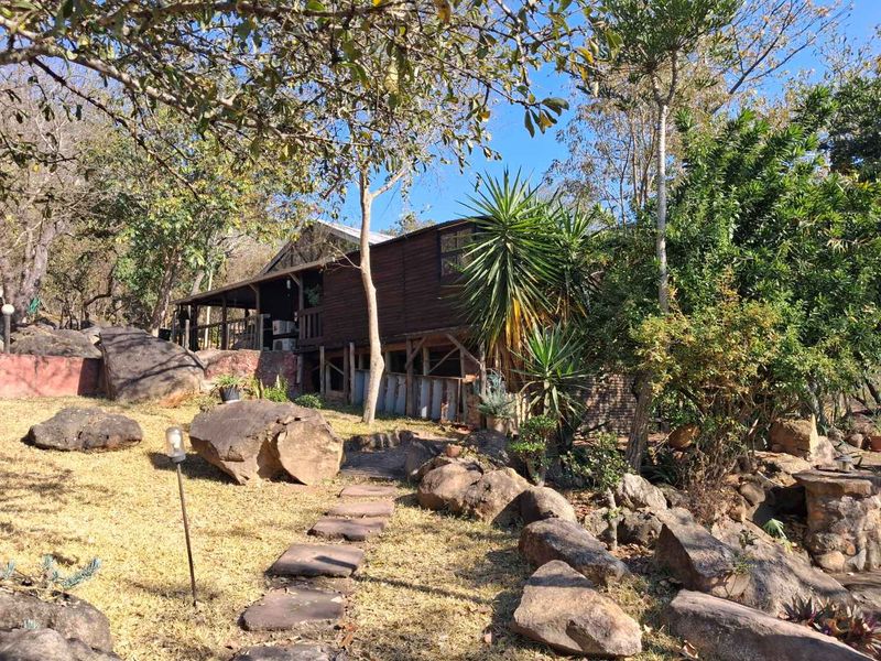 A rustic timber-clad dwelling set within a natural, rocky garden landscape featuring a raised wooden deck and stone pathway.