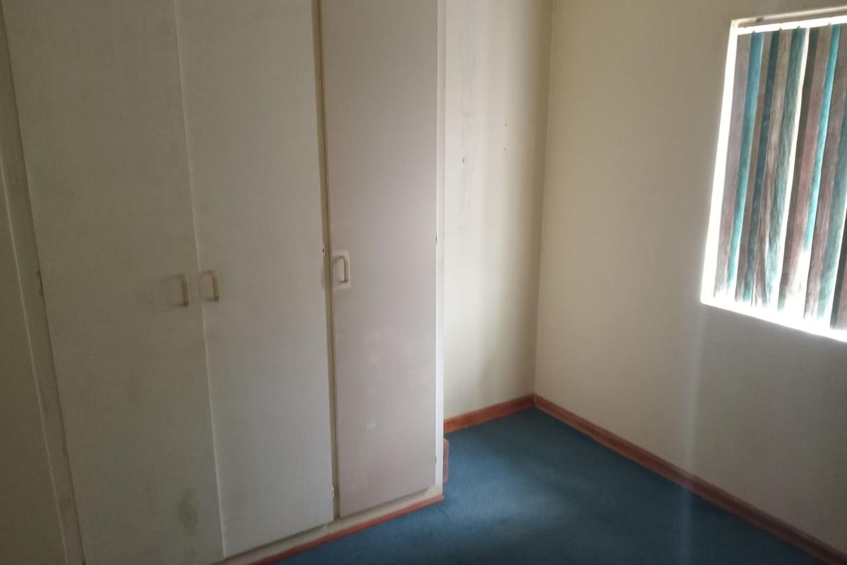 An empty bedroom featuring built-in cupboards, blue carpeting, and vertical window blinds. The room requires cosmetic updates to improve its overall condition.