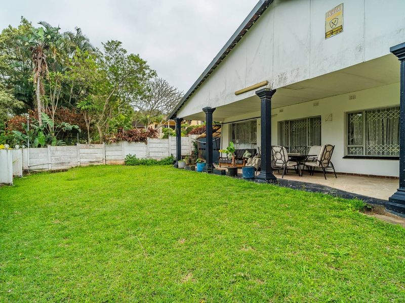 Rear view of the property featuring a covered patio area, tiled flooring, decorative columns, and a spacious lawn with a precast boundary wall.