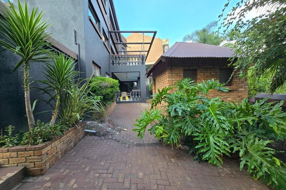 A rear exterior view featuring brick paving, landscaped planter boxes, a covered balcony, and a detached brick outbuilding.