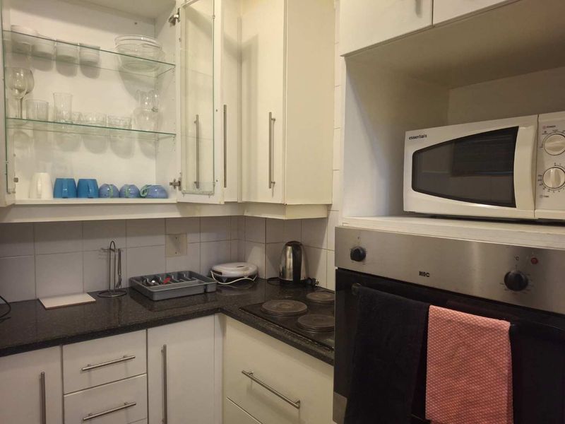 Compact kitchen featuring white cabinetry, dark granite countertops, built-in oven, electric hob, and tiled backsplash with integrated glass shelving.
