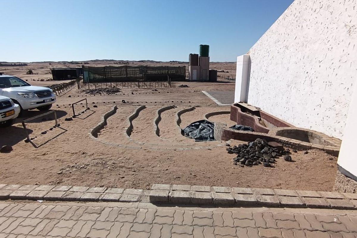 Exterior view of a small holding property featuring a large white wall, unfinished circular garden beds, and water storage tanks in the distance.